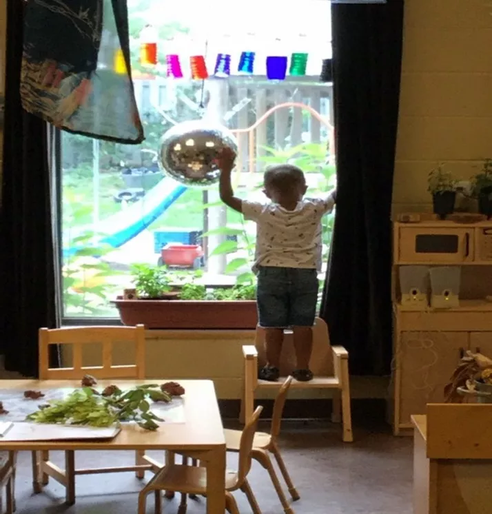 child-looking-out-window-connection ბავშვი დგას დიდ ფანჯარასთან და აკვირდება ეზოს, ბუნებრივი განათება ინტერიერში.